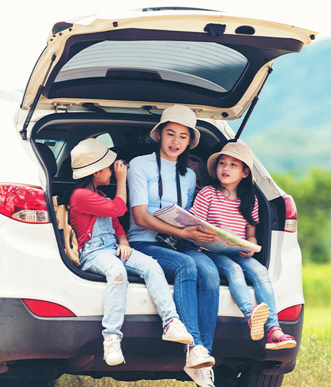 ラゲッジを開けた白いSUVの車内に座り、地図を見る大人と双眼鏡をのぞく子どもたち